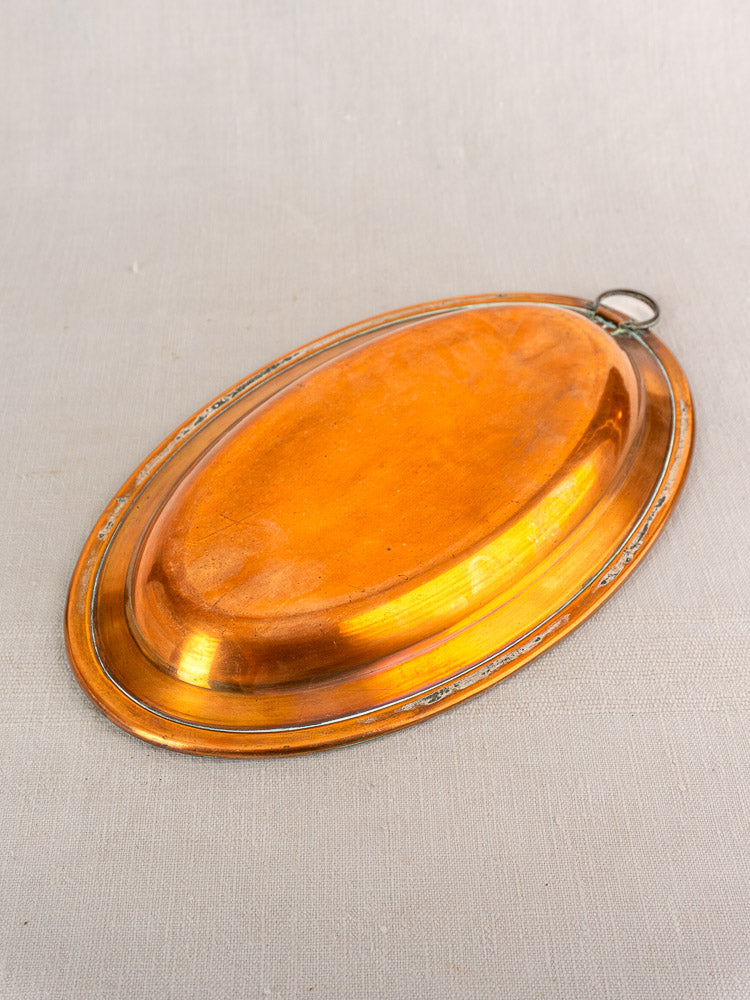 Antique French copper oval platter with tinned interior, decorative rim, raised center, and hanging ring on beige fabric background.