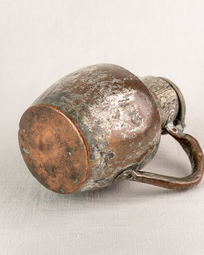 Antique French metal mug with worn handle, rounded base, on neutral fabric background.