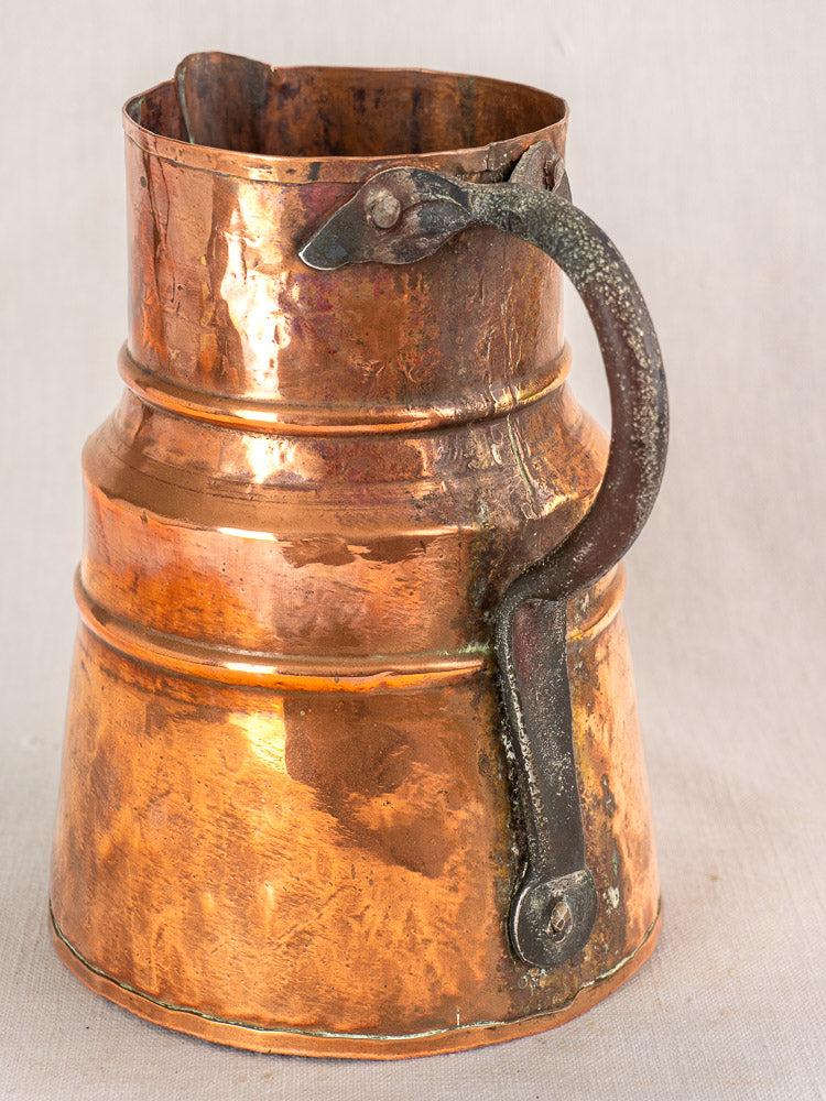 Antique French copper measure jug with forged iron handle, wide base, and tarnished finish on neutral fabric background.