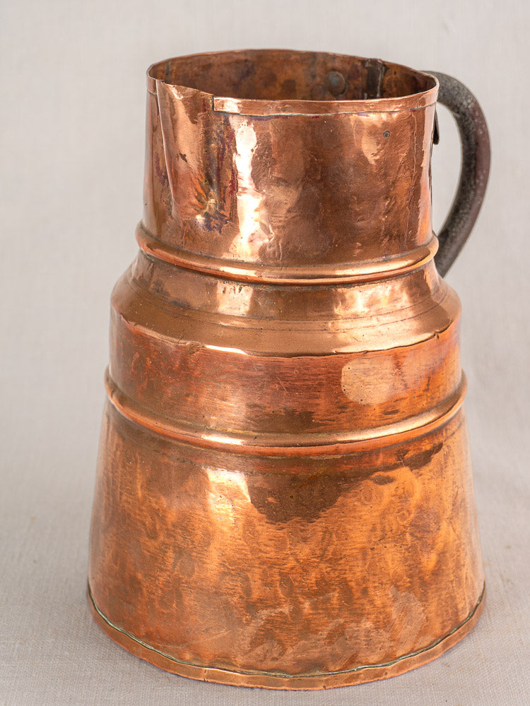 19th-century French copper measure jug with forged iron handle, wide base, and aged patina against a plain background.