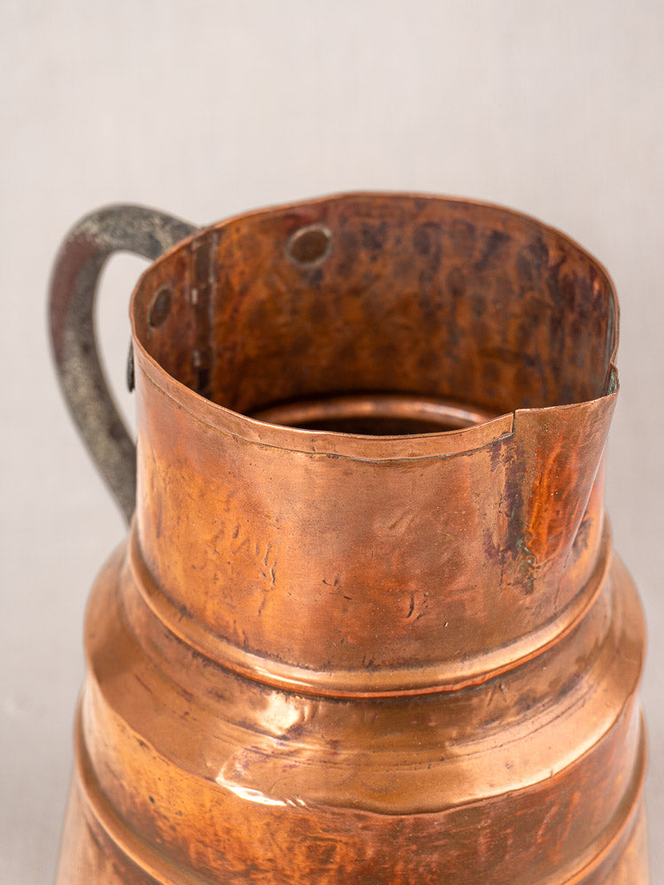 Antique French copper measure jug, hand-hammered, with forged iron handle, aged patina set against a neutral background.