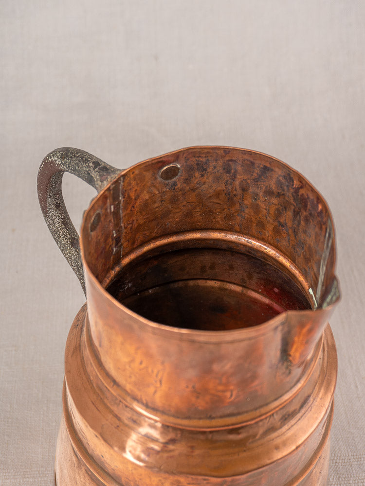 Antique French copper measure jug with forged iron handle, worn interior and spout on light fabric background.