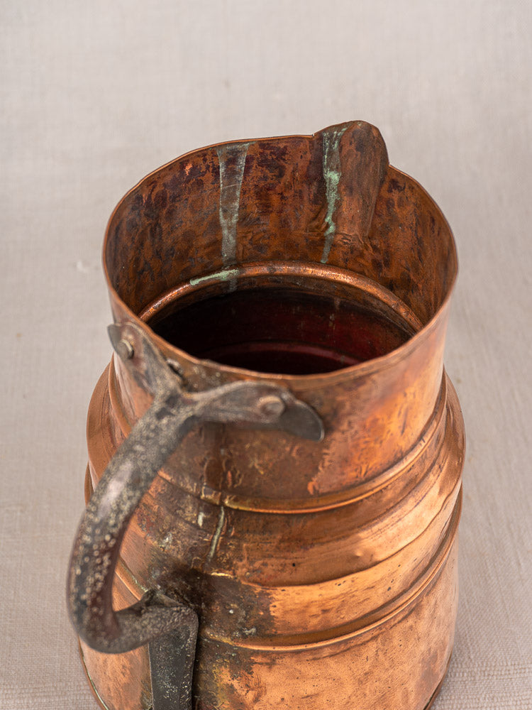 Antique French copper measure jug, hand-hammered with forged iron handle and wide rim, resting on light fabric.