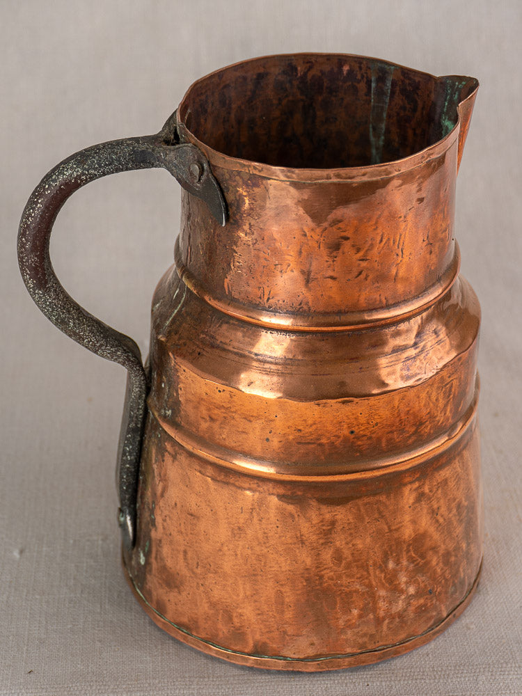 Antique French copper measure jug with hand-hammered finish and iron handle, aged patina on a neutral surface.