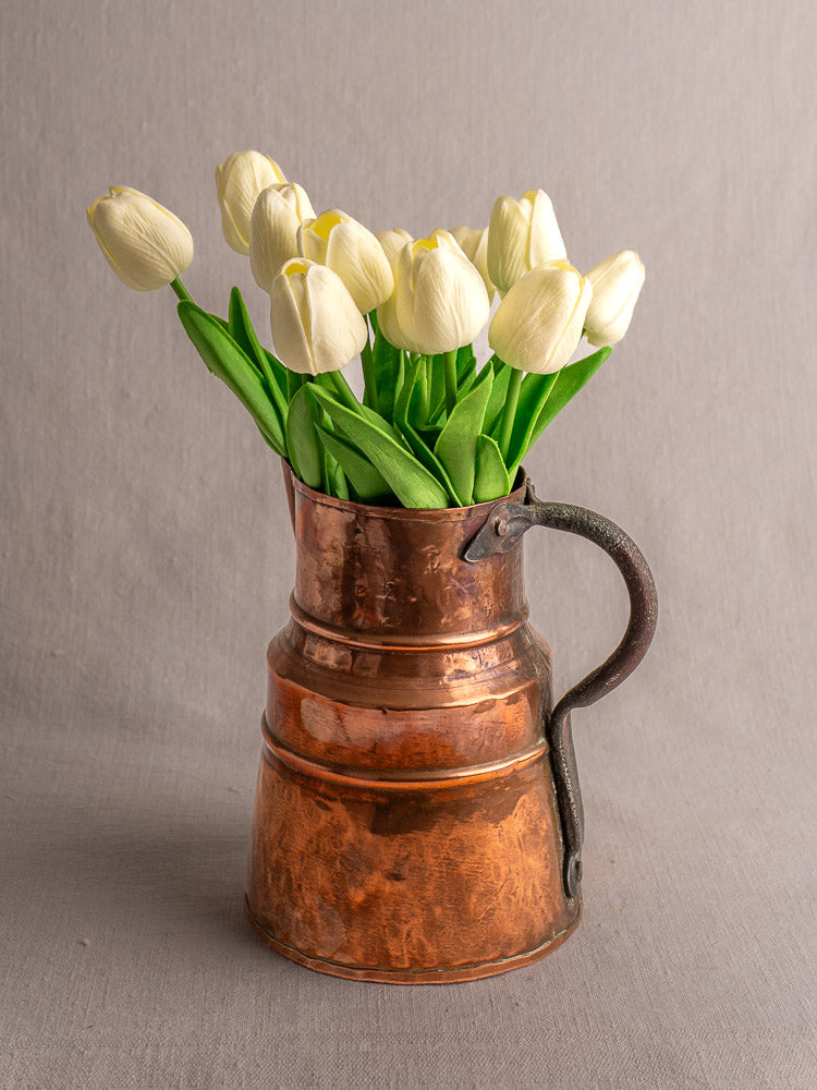 Antique French copper measure jug with forged iron handle, filled with white tulips, on a neutral gray background.
