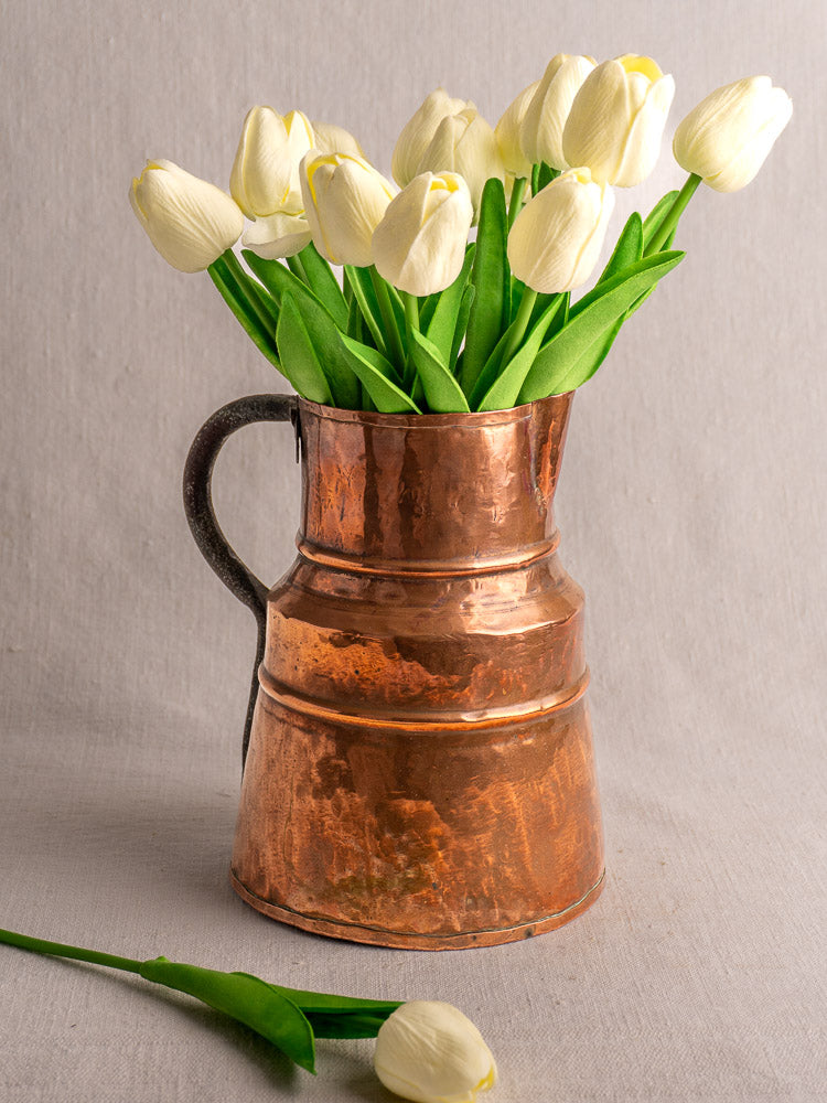 19th-century antique French copper measure jug with hand-hammered finish, filled with white tulips on light fabric.
