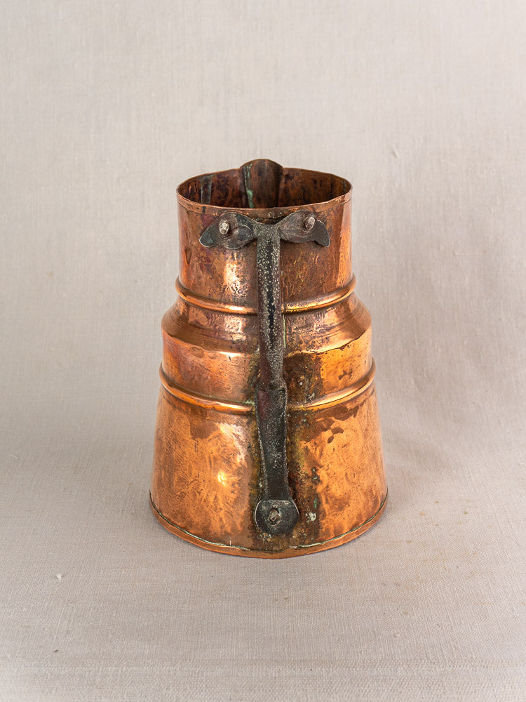 19th-century French antique copper measure jug, hand-hammered with forged iron handle, on plain light fabric background.