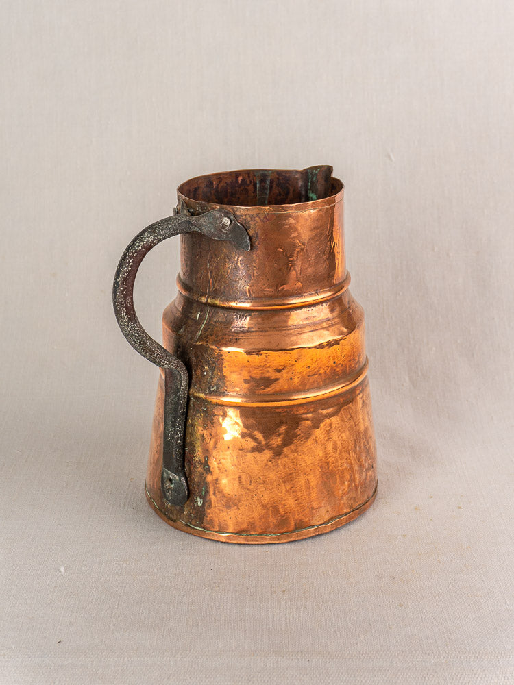 Antique French copper measure jug, 19th century, hand-hammered ribbed body, forged iron handle on beige fabric.