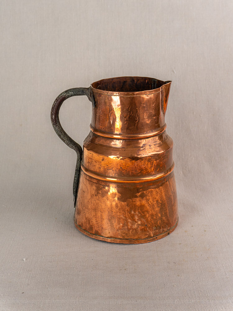 Antique French copper measure jug with forged iron handle on a light gray fabric background.
