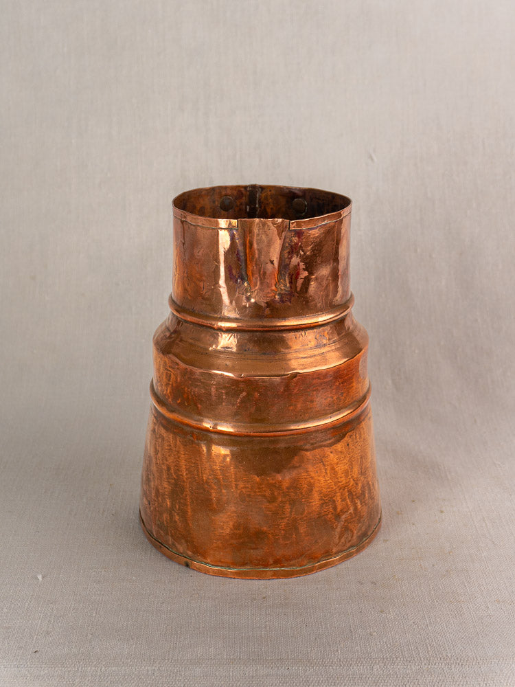 19th-century French hand-hammered copper measure jug with forged iron handle on a light fabric background.
