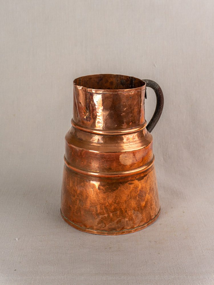 Antique French copper measure jug with forged iron handle and raised bands on a neutral fabric background.