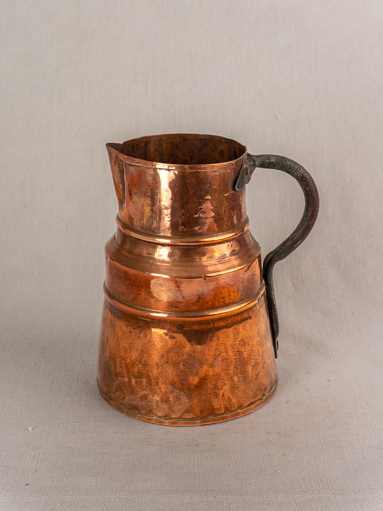 Antique French copper measure jug with forged iron handle on a light grey textured surface.
