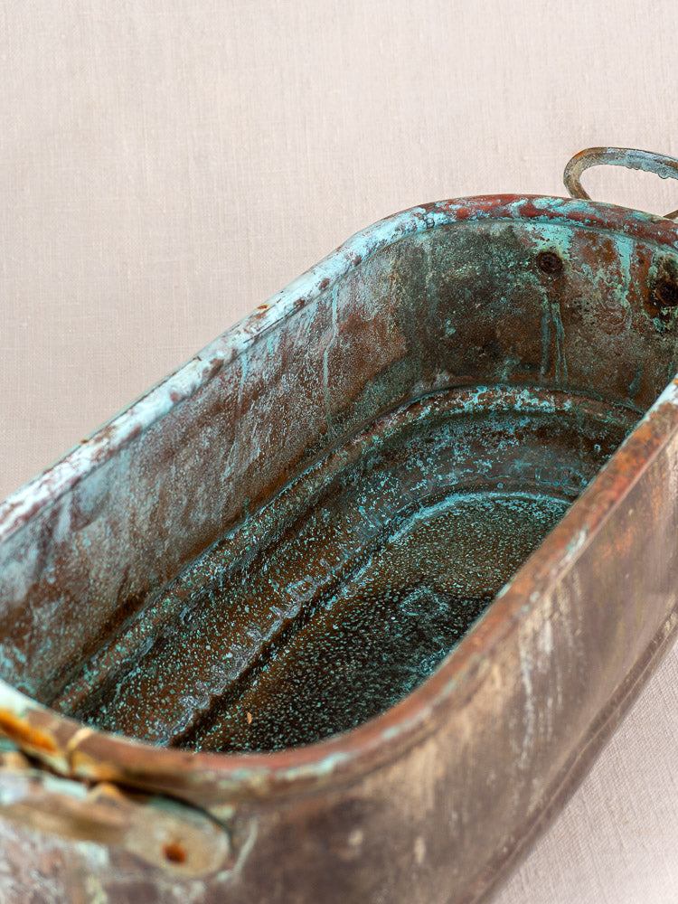 Antique French copper poissonnière jardinière with handles, verdigris patina, and visible tarnish on light fabric background.