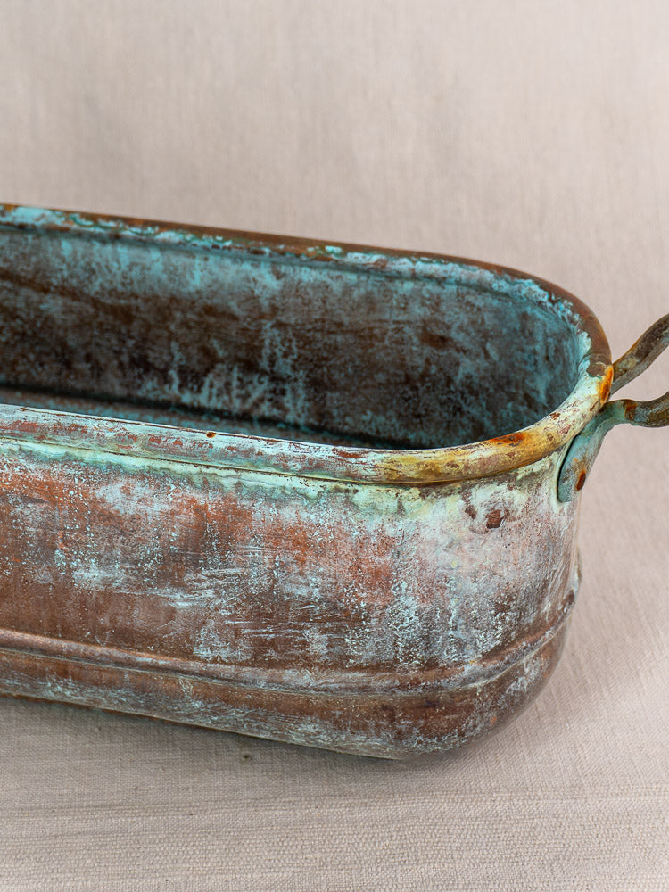 Antique French copper poissonnière jardinière with verdigris patina and corrosion, set on a plain light fabric background.