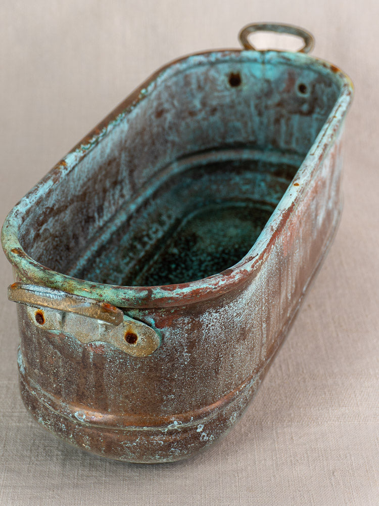 Antique French copper poissonnière jardinière with verdigris patina and handles on a light fabric surface.