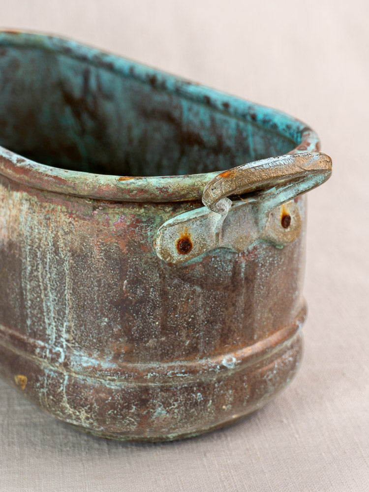 Antique French copper poissonnière or jardinière with verdigris patina and metal handle on beige fabric background.