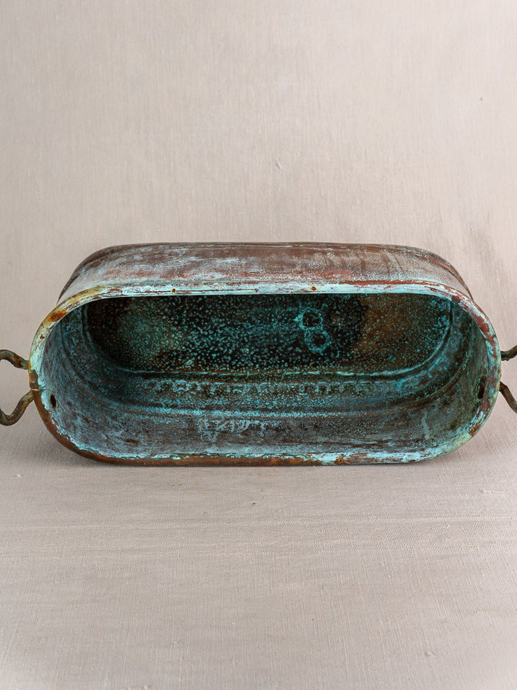 Antique French copper poissonnière jardinière with handles, verdigris patina, rusted edges on neutral fabric background.