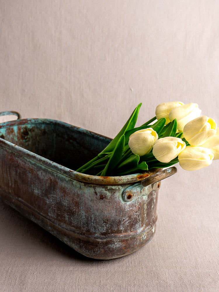 Antique French copper poissonnière jardinière with verdigris patina, holding pale yellow tulips and green leaves on fabric.