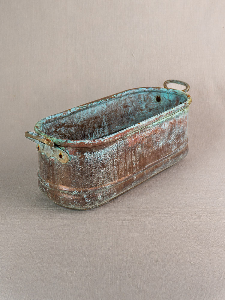 Antique French copper poissonnière jardinière with verdigris patina and brass handles on neutral fabric background.