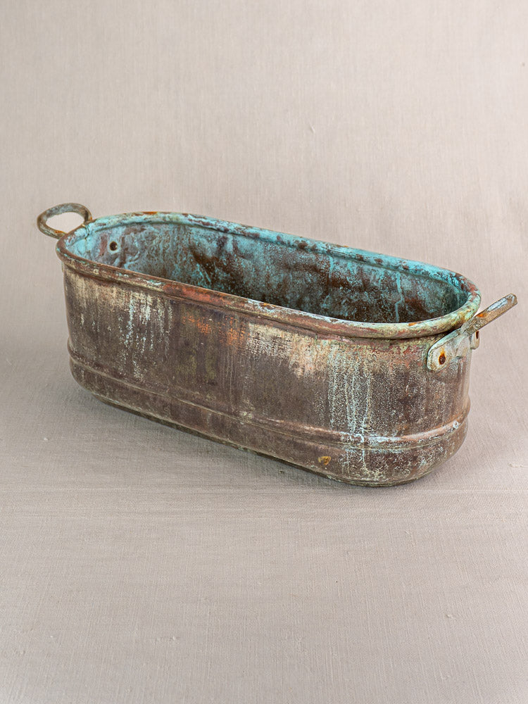 Antique French copper poissonnière jardinière with verdigris patina, oval and double-handled, on beige background.
