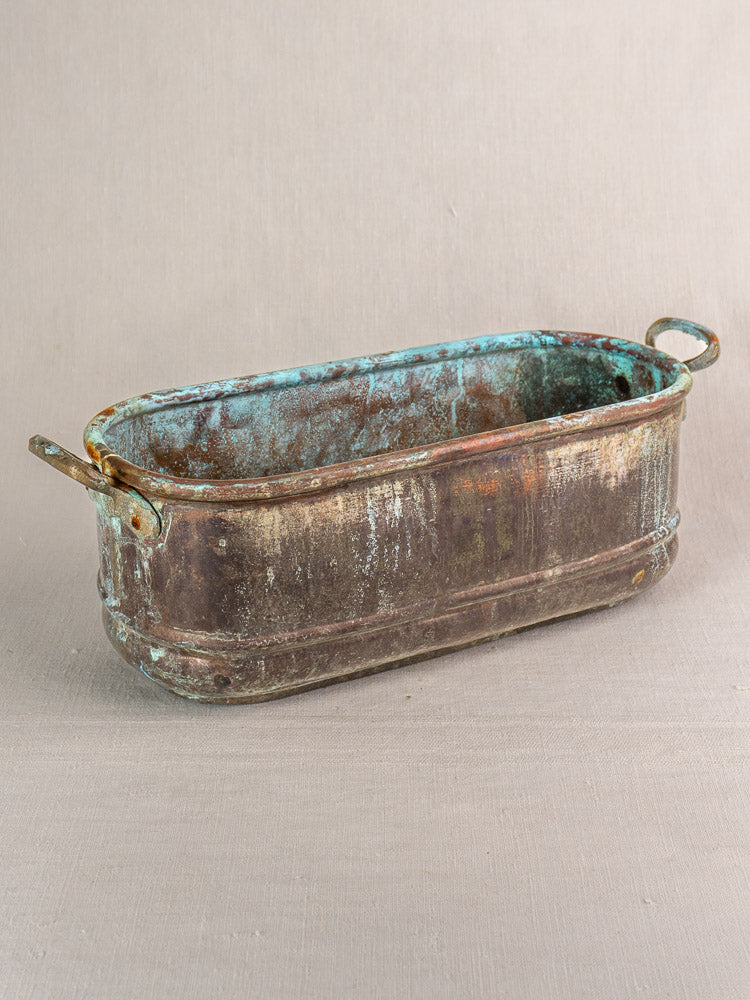 Antique French copper poissonnière jardinière with verdigris patina and dual handles on neutral fabric background.