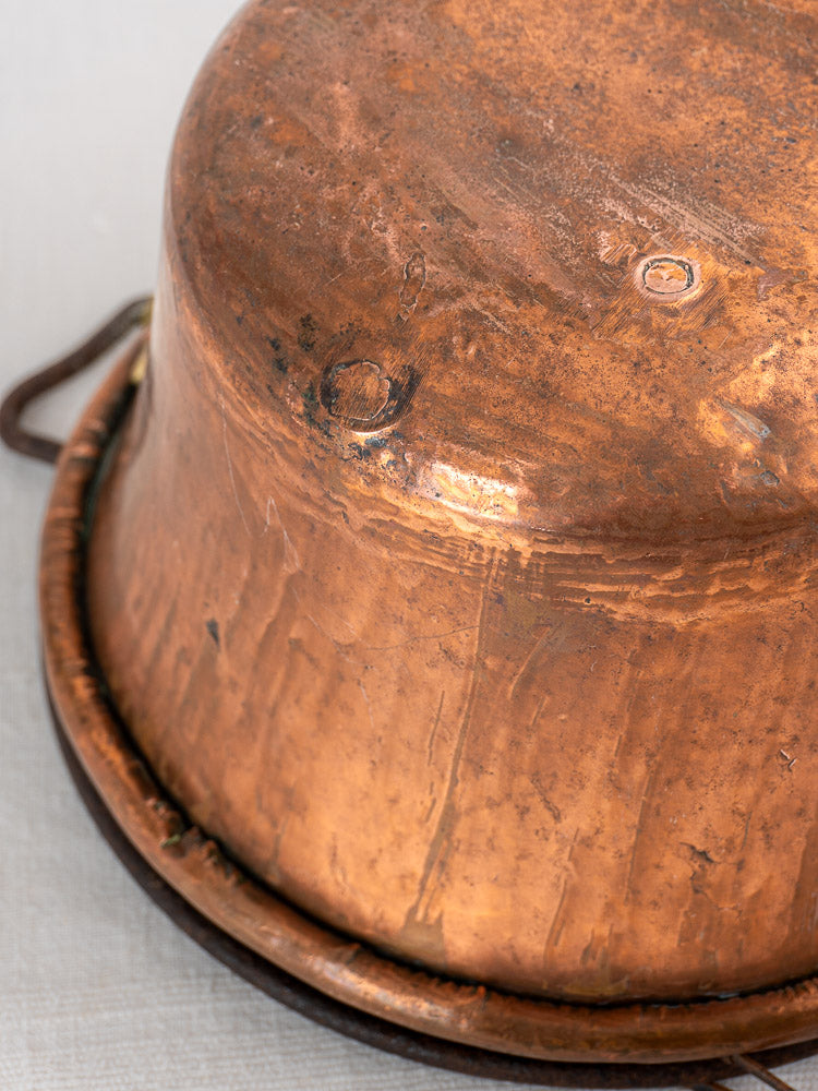Antique French copper hearth pot upside-down, iron bail handle and chain, patina and dents visible.