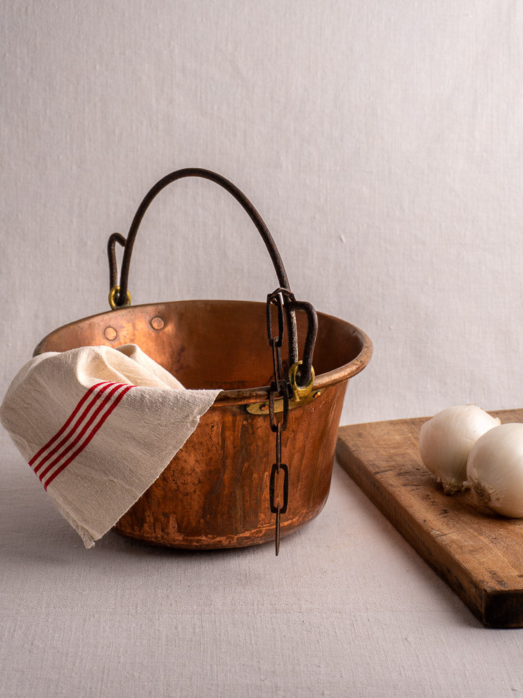 Antique French copper hearth pot with iron bail handle and chain on white surface, striped cloth draped, onions beside.