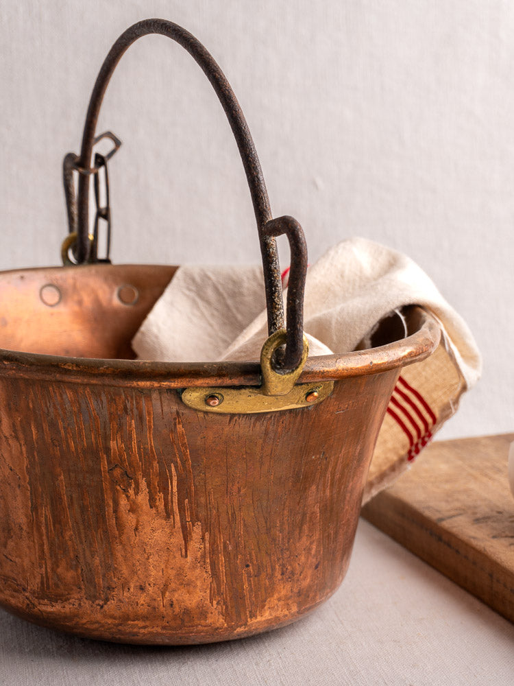 Antique French copper hearth pot with iron bail handle and chain on wood and striped cloth.