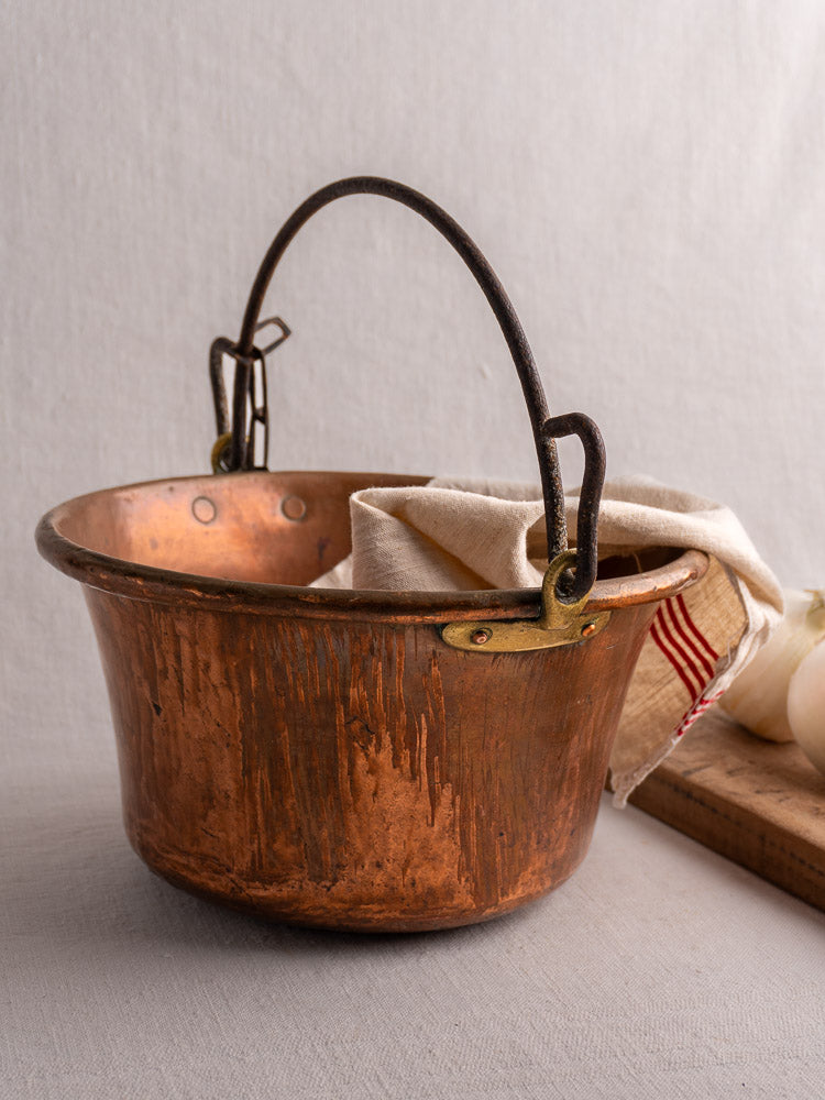 Antique French copper hearth pot with iron handle and chain, holding a striped beige cloth, next to white onions and a cutting board.