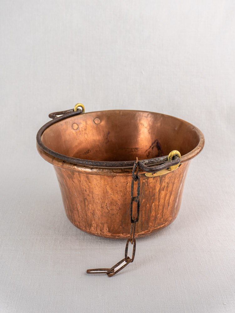 Antique French copper hearth pot with iron bail handle and chain on light fabric surface.
