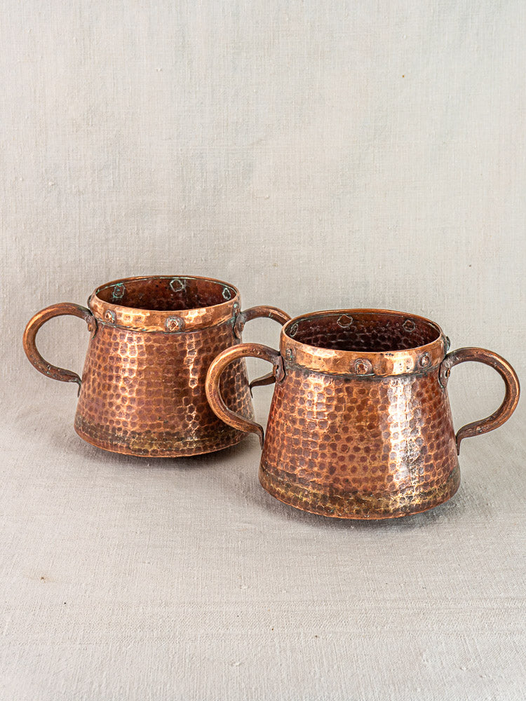 Pair of antique French copper pots with rivet accents and three handles on a neutral fabric backdrop.