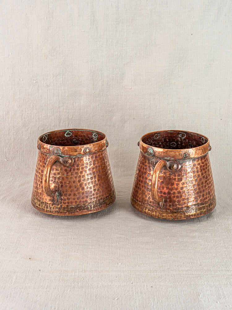Pair of antique French hand-hammered copper pots with round bases and curved handles on neutral fabric background.
