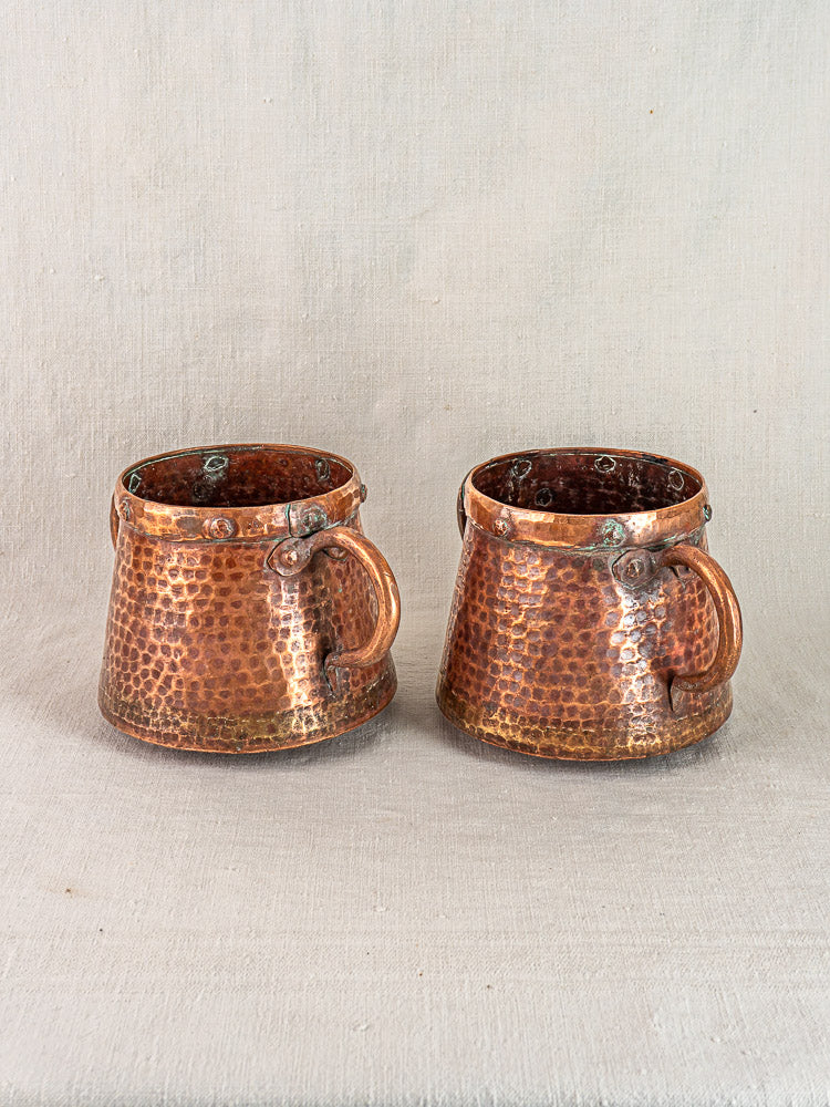 Pair of antique French hand-hammered copper pots, circa 1880–1920, on soft light fabric background.