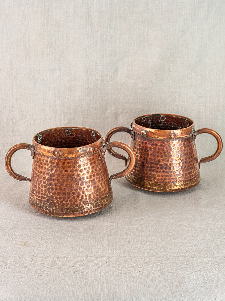 Pair of antique French hand-hammered copper pots from 1880–1920, shown side by side on neutral fabric.