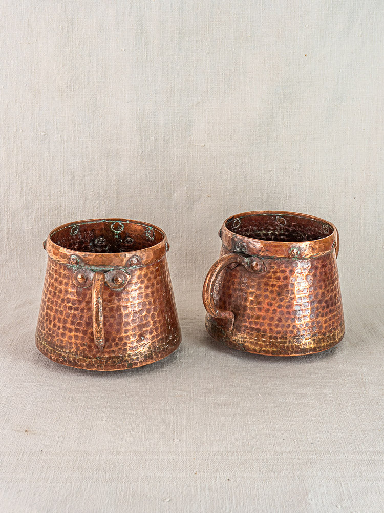 Pair of antique French hand-hammered copper pots with patina and sturdy handles on neutral fabric background.