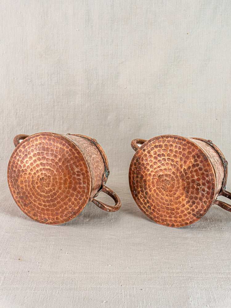 Pair of antique French copper pots, hand-hammered with textured bottoms and side handles, on neutral fabric background.