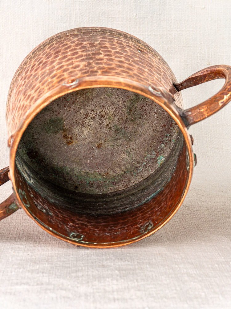 Antique French copper pot with hand-hammered finish, two handles, and oxidized interior on light fabric background.