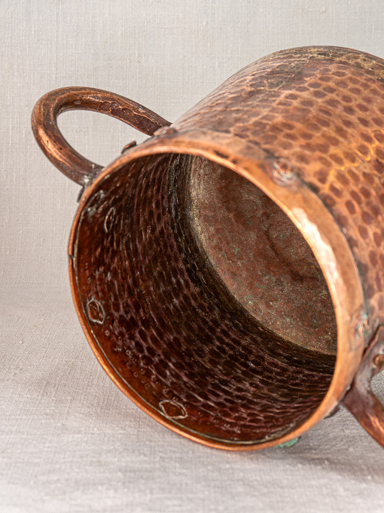 Antique French copper pot with two handles, hand-hammered, showing textured interior on light fabric surface.