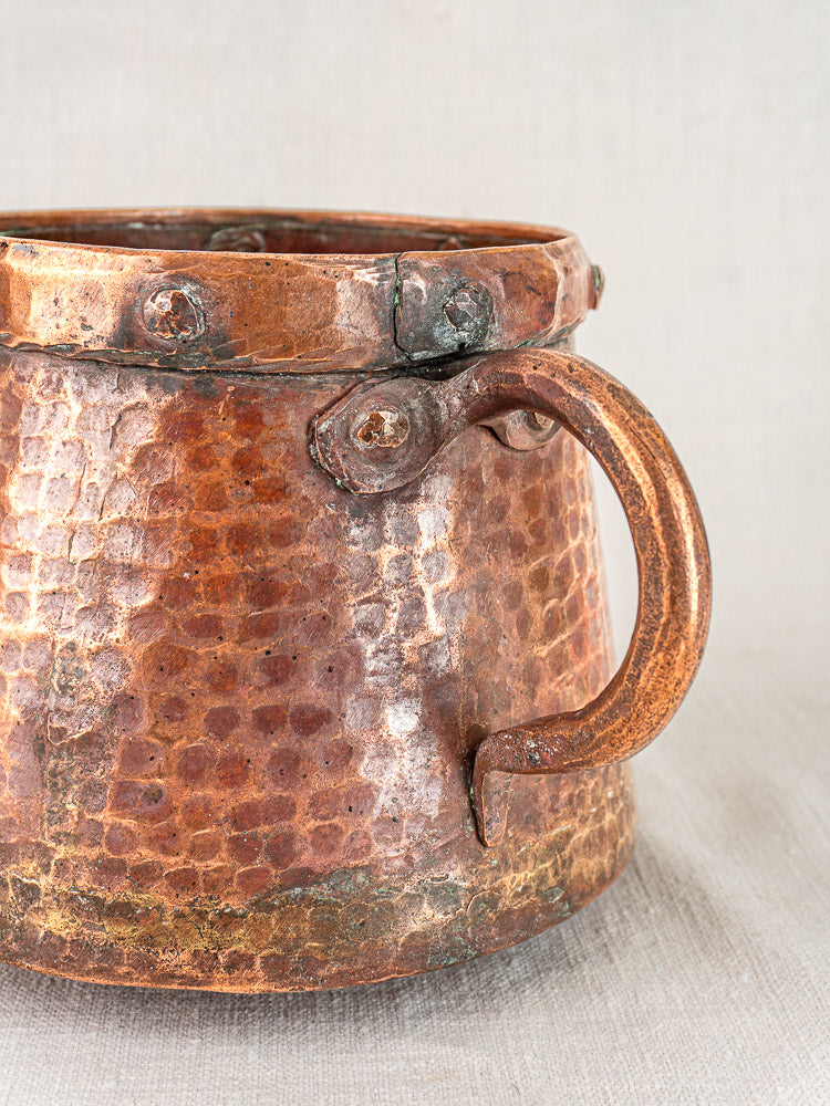 Antique French hand-hammered copper pots pair with aged patina and riveted handles on neutral fabric background.
