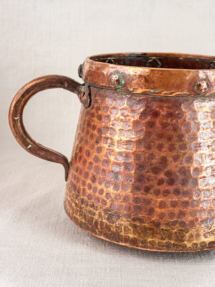 Hand-hammered French antique copper pots with curved handles and rivets on light textured fabric background.