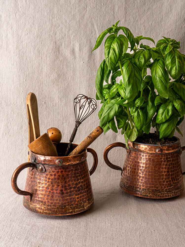 Pair of antique French copper pots with handles, one holding utensils, the other fresh basil, on neutral fabric.