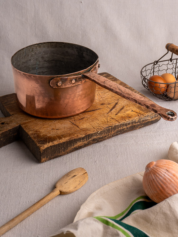 18th-century French copper preserving pot with solid handle on wood board, near spoon, striped cloth, garlic, eggs in basket.