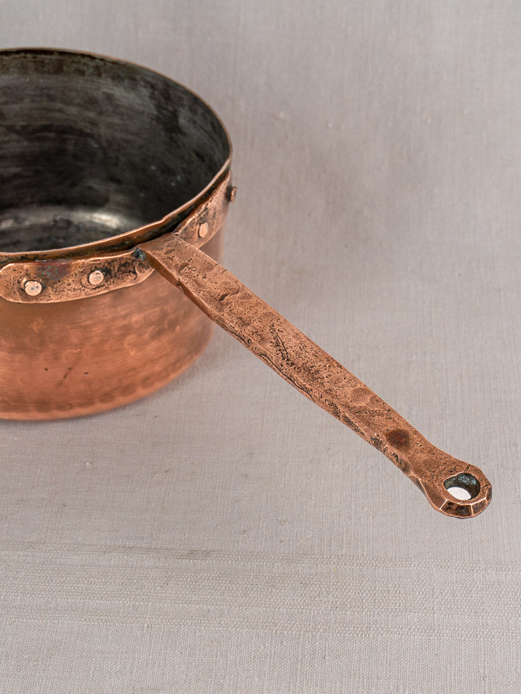 Antique French copper preserving pot with hammered finish, solid handle, and hand-forged rivets on a light fabric surface.
