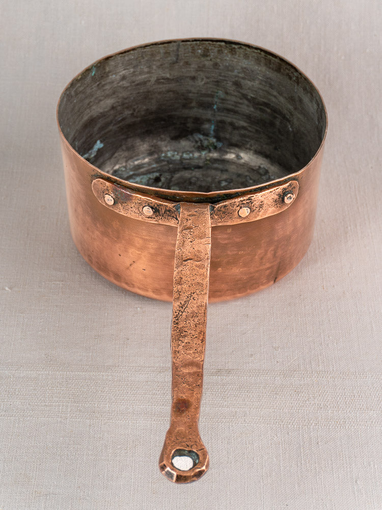 Antique French copper preserving pot with solid handle, hand-forged rivets, and patina, on a light fabric surface.