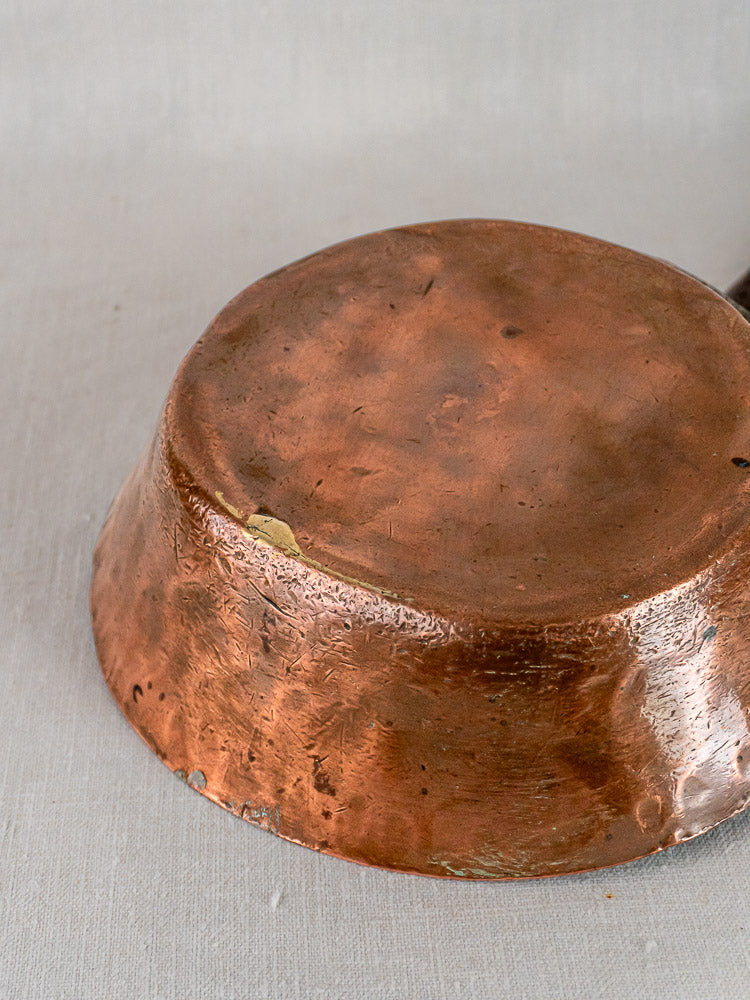Bottom view of antique French copper sauté pan with wear, dents, and chipped edge on gray surface.