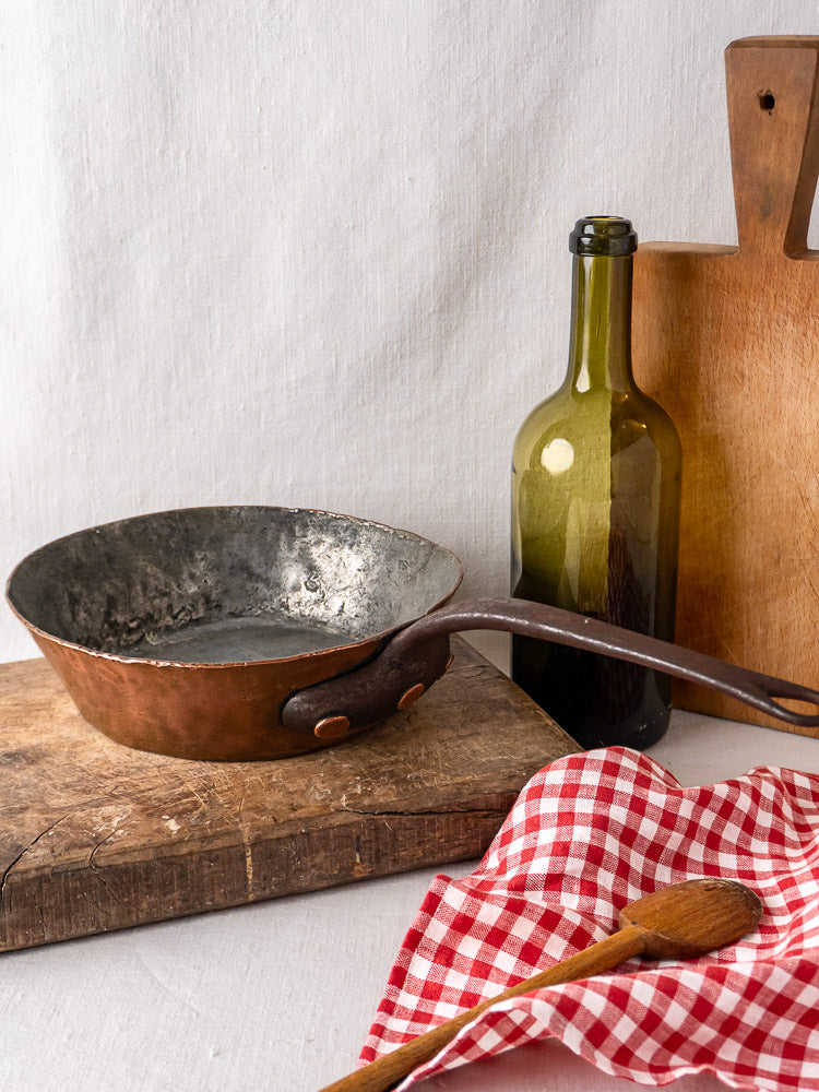 Antique French copper sauté pan on wood surface with wine bottle, boards, spoon, and checkered cloth.