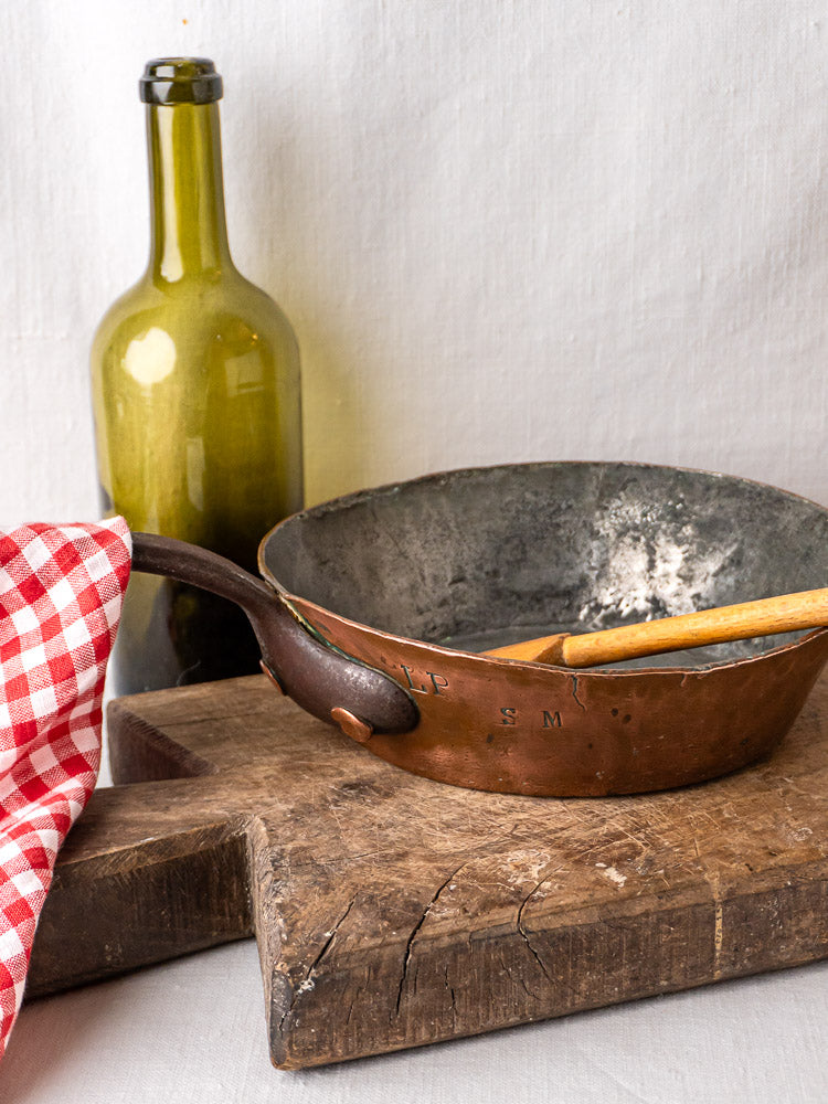 French copper sauté pan with wooden spoon and green glass bottle on rustic cutting board.