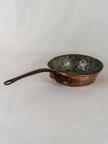 Antique 19th-century French copper sauté pan with dark handle on light fabric background, hand-hammered patina.