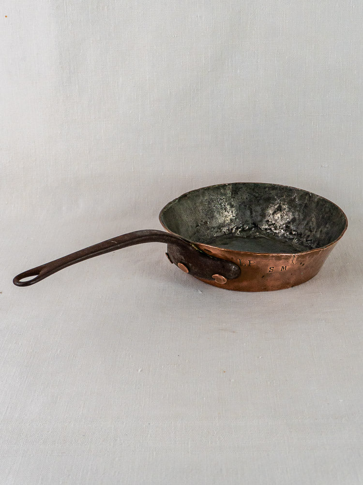 Antique 19th-century French copper sauté pan with dark handle on light fabric background, hand-hammered patina.