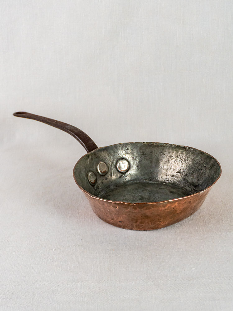 Antique 19th-century French copper sauté pan with hand-hammered body and dark metal handle on light surface.